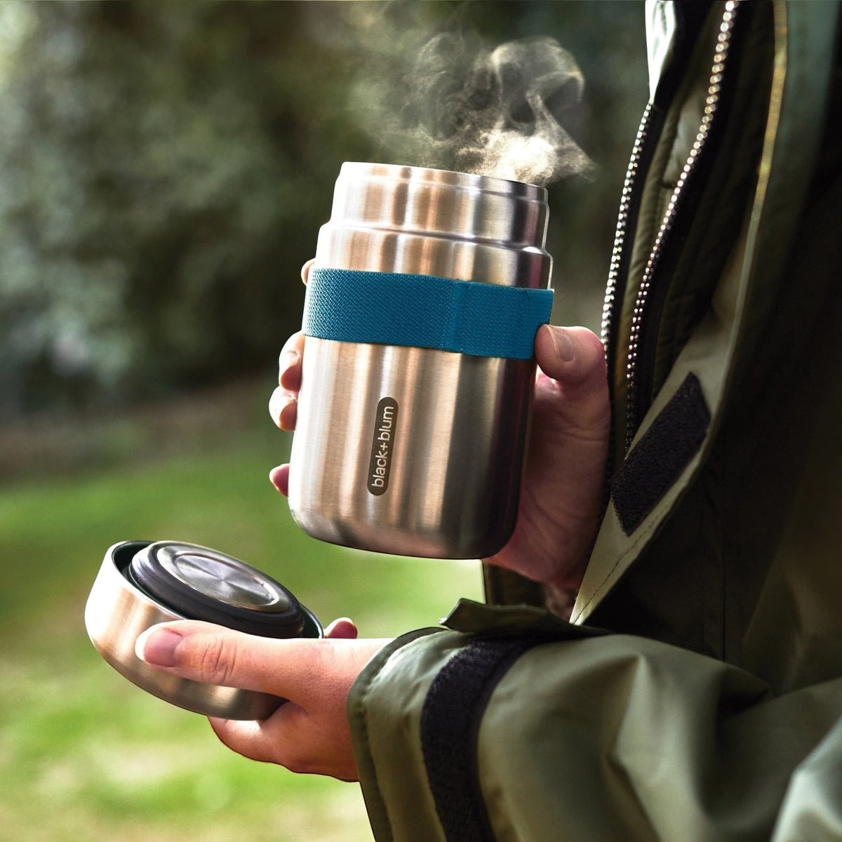 Lunchpot Black+Blum argenté tenu dehors, couvercle séparé et vapeur visible